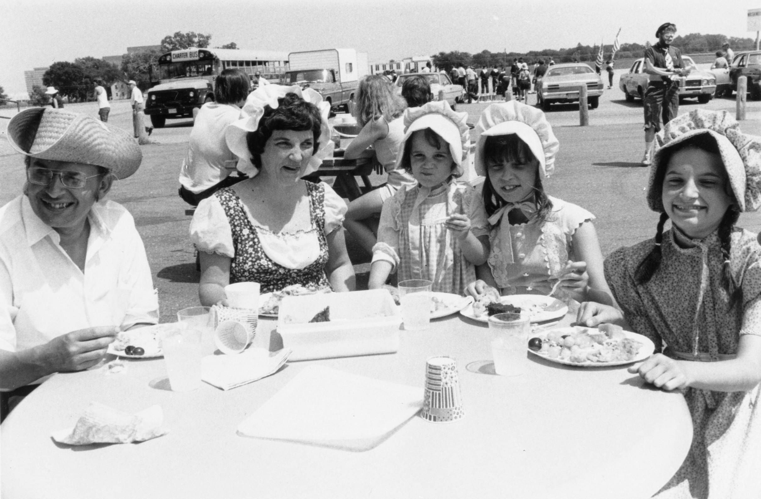 Black and white photo of five people seated at a picnic table outdoors, enjoying a meal. Four are wearing pioneer-style bonnets and dresses, while one man wears glasses and a straw hat. Vehicles and people are visible in the background.